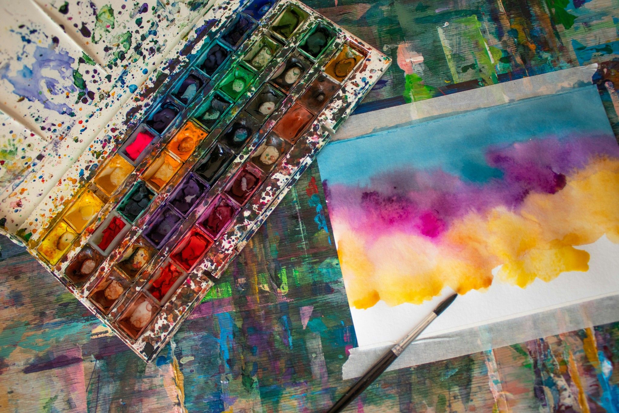 Watercolor paint palette, brush and colorful wash painting on a paint-spattered table.